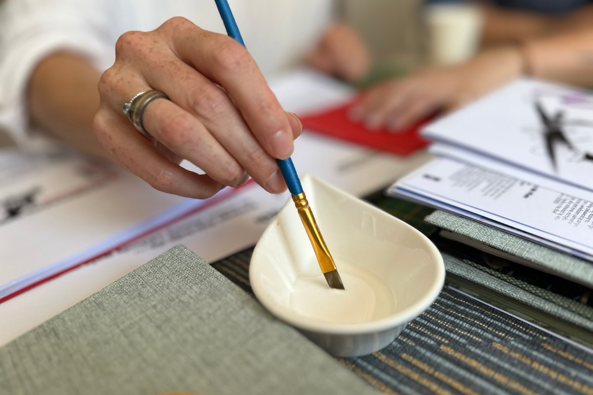 Na zdjęciu widać rękę z pędzelkiem zanurzającą się w miseczce z klejem - praca introligatorska podczas własnoręcznego wykonywania notesu.