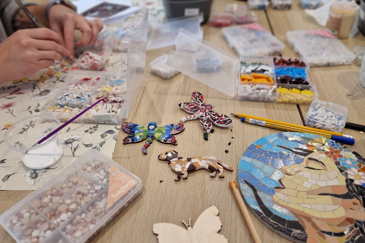 warsztaty mozaikowe dla dorosłych w poznaniu. na zdjęciu widać mozaikę przedstawiającą ciemnowłosą kobiete z zamkniętymi oczami i wianku na głowie. Na środku mozaikowe broszki: ważki i pies jamnik. Widać też ręce przy pracy oraz materiały