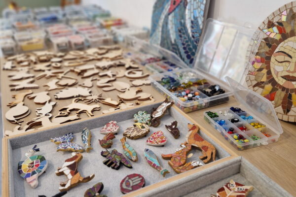 mozaikowe broszki i magnesy wykonane z kawałków ręcznie ciętego szkła i ceramiki . Na drugim planie drewniane bazy, które można wykorzystać do stworzenia swojego unikatowego projektu. W tle duże mozaiki wykonane przez Zbyszka z Tesspiry