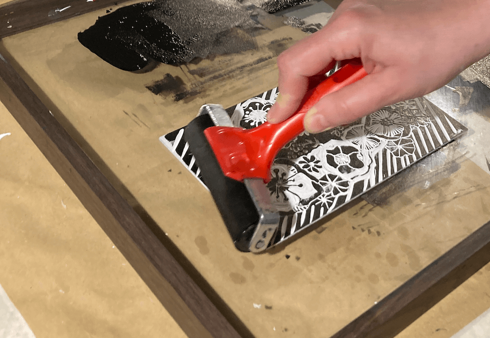 Na zdjęciu widać wyłaniającą się spod wałka czarno-białą matrycę linorytu. WIdać linie tła, kwiaty. Matryca leży na przezroczystej szybie.